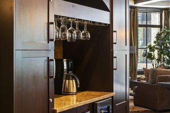 A built-in wet bar area with dark wooden cabinets, a granite countertop, and glassware hanging above, next to a mini-fridge.