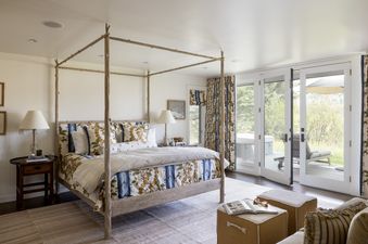 A serene and elegantly decorated master bedroom at the Retreat on the Creek, featuring a natural wood four-poster bed with patterned bedding, flanked by nightstands with lamps, and large glass doors opening to an outdoor deck with a hot tub and scenic vie