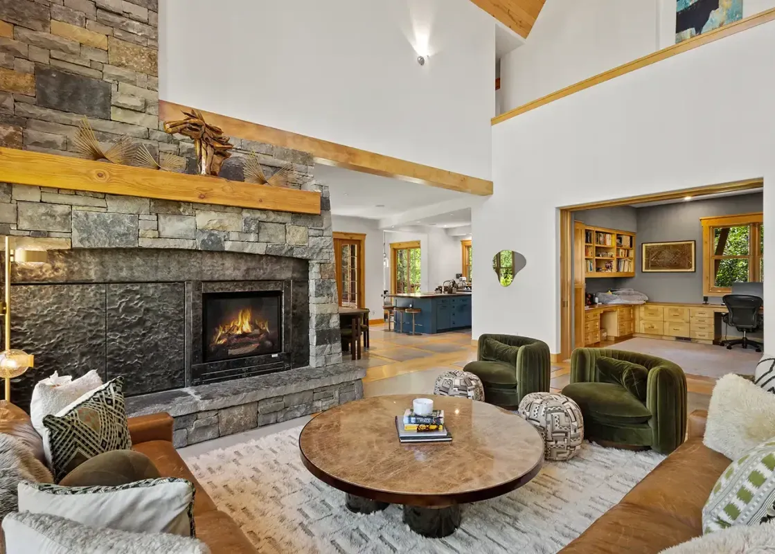 A spacious and inviting great room with a large stone fireplace, comfortable seating, and views into the kitchen and a home office.