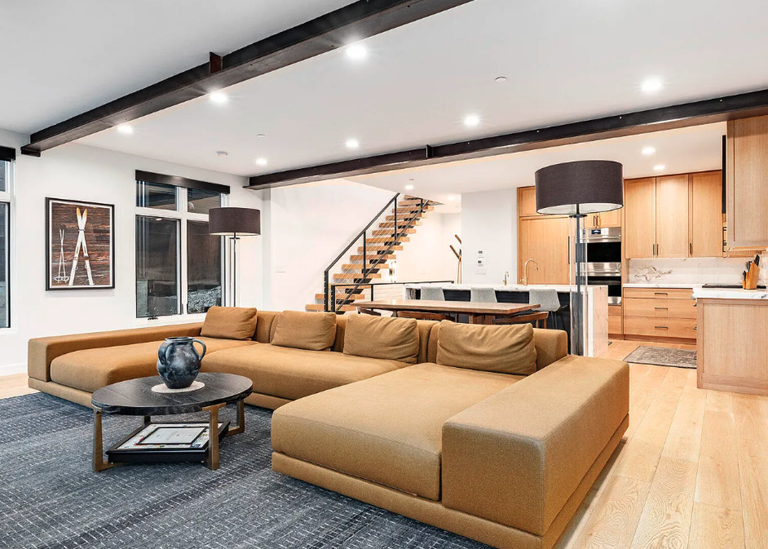 A contemporary open-plan living area at Kings Crown Hideaway, featuring an L-shaped sectional sofa, modern coffee table, and an integrated kitchen and dining area, all under a ceiling with exposed dark beams and recessed lighting.
