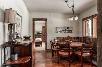 A cozy dining area features a round wooden table and chairs, illuminated by a chandelier, with a rustic wainscoting and a window.