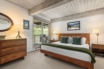 A serene bedroom at Enclave 211 features a comfortable bed with a patterned duvet and green accents, rich wooden furniture, elegant lighting, and direct access to a balcony with views of green landscapes.