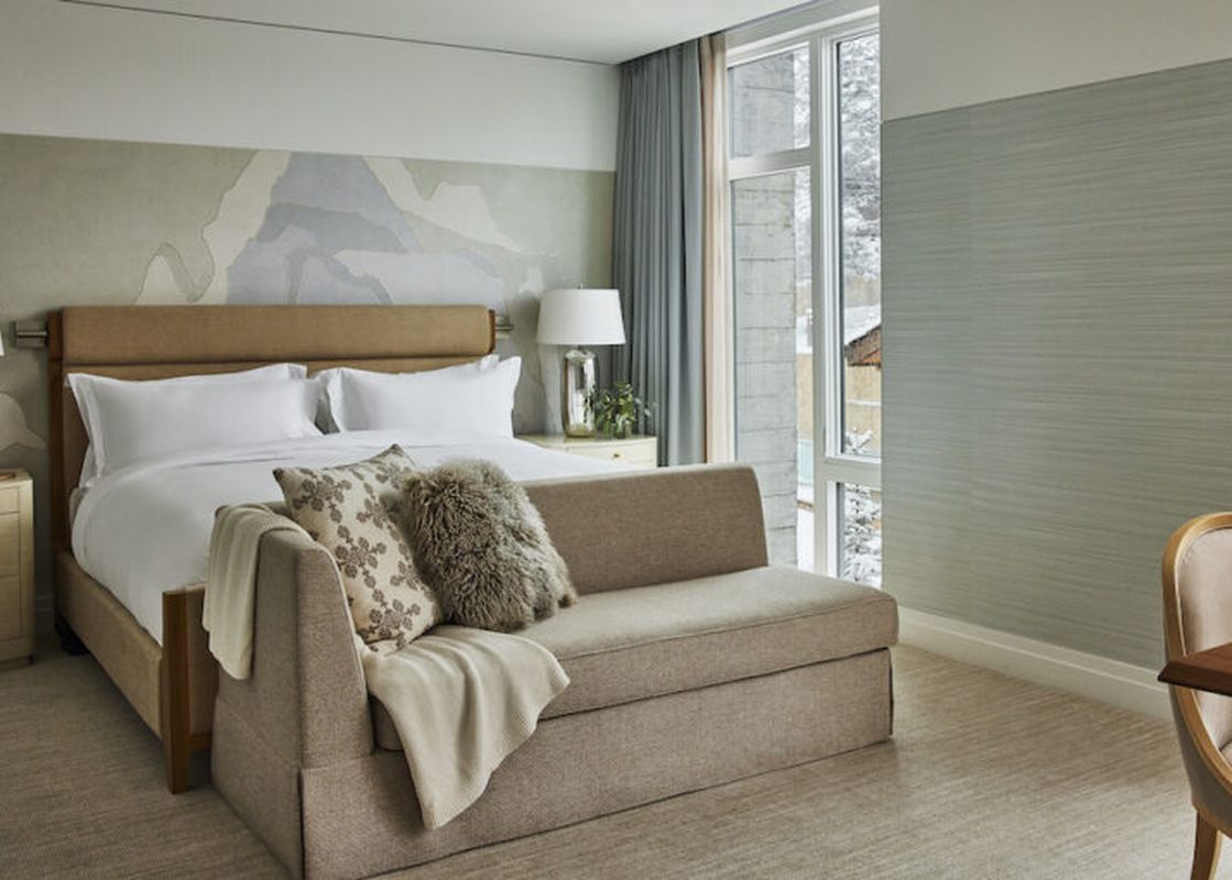 A modern hotel bedroom with a king-size bed, a couch, a table and a chair, and a window with a view of snow-covered trees.