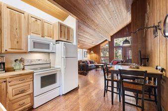 An open-concept living area with a kitchen, dining table, and living room featuring large windows and wooden vaulted ceilings.