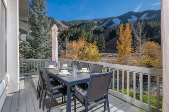 An outdoor dining table set for eight on a balcony overlooks a scenic mountain range with fall foliage and a river.