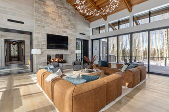 A spacious living room at Lone Star Lodge Mountain Village features a large brown sectional sofa, a stone fireplace, a flat-screen TV, and expansive windows offering a winter mountain view.