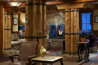 A warmly lit living room at The Inn at Lost Creek Junior Suite features robust wooden columns, a cozy stone fireplace, comfortable seating, and elegant floral decorations, creating an inviting rustic-chic atmosphere.