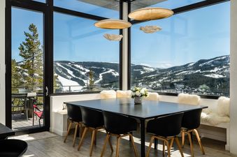 A modern dining area with a black table and chairs, featuring floor-to-ceiling windows that offer a panoramic view of snow-covered mountains and pine trees.