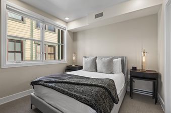 A modern bedroom in Rio Grande 301 with a grey upholstered bed, two dark wood nightstands with lamps, and a large window overlooking an adjacent building.