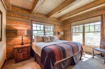 A cozy bedroom with rustic wooden walls and ceiling, featuring a queen-sized bed with a striped throw blanket, two nightstands with lamps, and large windows looking out to a sunny outdoor scene.