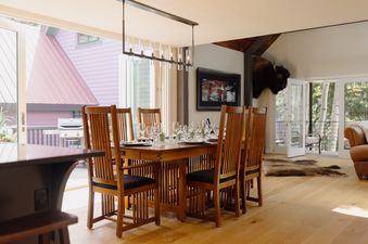 A beautifully set wooden dining table with chairs, wine glasses, and floral arrangements, illuminated by a modern chandelier, with large sliding glass doors revealing a vibrant pink building and lush greenery outside.