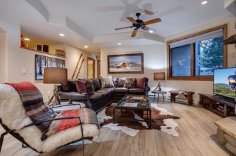 A cozy lounge area with a dark leather sectional sofa, a cowhide rug, and ski-themed decor, illuminated by lamps and natural light from a large window.