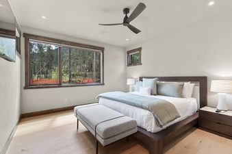 A serene bedroom at Independence Lodge features a large bed with white and light blue bedding, two bedside lamps, a gray bench, and a large window overlooking a forest.