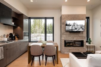 The kitchen and living area of 230 LakeLodge, showcasing modern design, a dining table, and a stone fireplace with a TV and a view of snow-dusted trees outside the window.