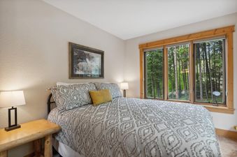 A cozy bedroom at Elk Run features a queen bed with patterned bedding, a wooden nightstand, and a large window overlooking a lush forest.