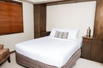 The comfortable bedroom at Landmark 705 features a queen-size bed, elegant wooden blinds, and modern built-in cabinetry with integrated lighting.