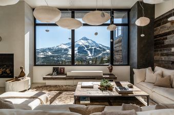 A luxurious and cozy living room featuring a large window with a panoramic view of snow-capped mountains and ski slopes, accented by unique pendant lights and rustic-chic furnishings.