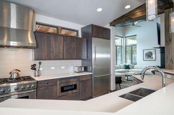 A modern kitchen with dark wood cabinets, white countertops, and stainless steel appliances, featuring a large refrigerator, gas range, and a dual-basin sink.
