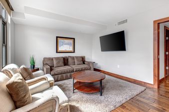 A cozy living room in Lowell 402A with a brown sofa, two armchairs, a round coffee table, and a wall-mounted TV.