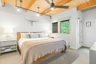 A primary bedroom with a king-sized bed, wooden ceiling, modern lighting, and a ceiling fan. The room has a unique silver bull skull wall decor, a stylish nightstand with a lamp, and a large window offering a view of lush greenery.
