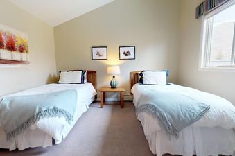 A guest bedroom at Westwind 402 features two twin beds with white bedding, a wooden nightstand with a lamp, and a large window offering mountain views.