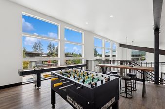 A bright games room with a foosball table, shuffleboard, and large windows offering views of the outdoors.