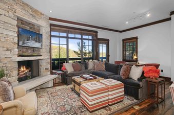 The Arrowleaf Lodge 205 living area features a stone fireplace, a large sectional sofa with colorful pillows, and expansive windows overlooking a mountain landscape.