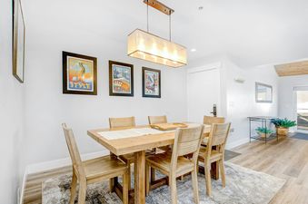 The dining area of Chatham Crossing 2198 features a wooden table and chairs with a large, bright window that allows for natural light. The walls are white and decorated with framed artwork that features the local landscape.