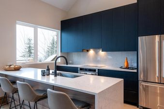 A modern kitchen at Northern Lights 32 featuring dark cabinetry, stainless steel appliances, a white island with bar seating, and a large window overlooking a snowy landscape.