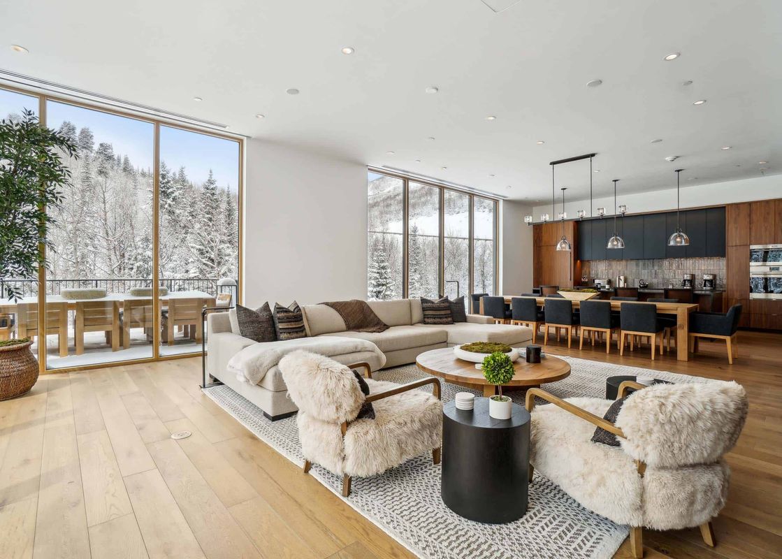 The spacious great room at Founders Place 1701 features a cozy living area with a sectional sofa and fur-upholstered chairs, alongside a dining area and kitchen, all framed by large windows revealing a snowy mountain landscape.