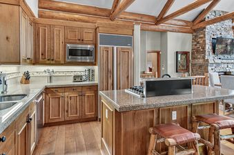 The kitchen at Timber Haven showcases rustic elegance with wooden cabinetry, a large kitchen island with a cooktop, and granite countertops, complemented by a stone fireplace in the adjacent living area.