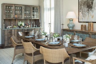 A rustic yet elegant dining area features a long wooden table set with blue napkins and wicker chairs, complemented by a wet bar and a decorative console table.