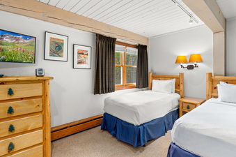 A cozy bedroom with two twin beds, rustic wooden furniture, framed artwork, a television, and a window with green outdoor views at Top of the Village Leaf 401.