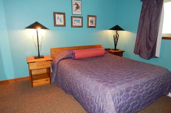 A cozy bedroom with a large bed, two bedside tables with lamps, and framed pictures on the teal wall.