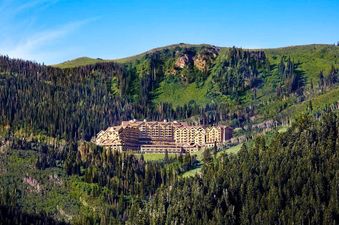 The Montage Deer Valley King Room Resort View is nestled within a lush mountain landscape under a clear blue sky, featuring a grand hotel complex surrounded by dense forests.