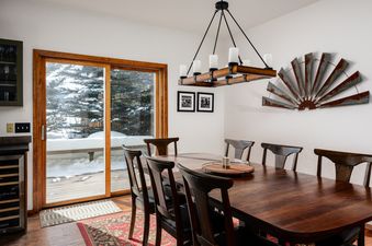 A rustic dining area with a dark wood table and chairs, illuminated by a chandelier, features a large sliding glass door opening to a snow-covered deck with winter tree views.