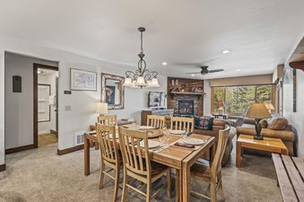 The dining area at Snow Flower 56 Park City features a wooden dining table, a chandelier, and an open-concept layout leading to a living room with a fireplace and large window.