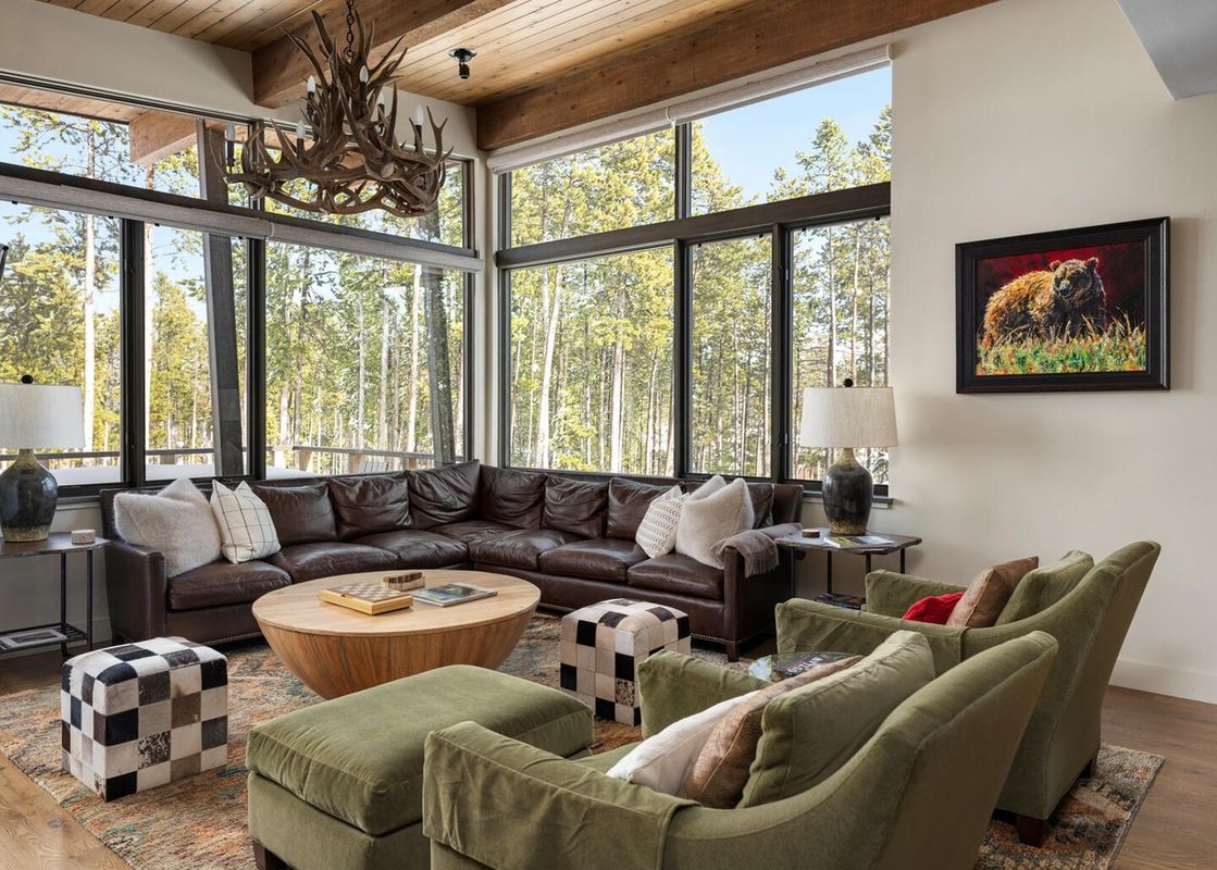 A rustic yet luxurious living room with a large leather sectional, round coffee table, and forest views through expansive windows.