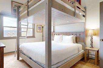A cozy bedroom with a wooden bunk bed, featuring a queen-sized lower bunk with white bedding and a star-adorned headboard, beside a window with an exterior view.