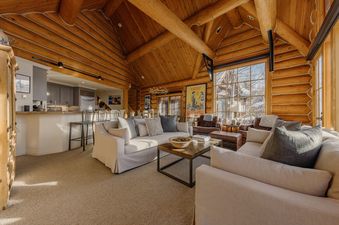 The living area of Frostpine Cabin at See Forever, featuring exposed log beams, cozy seating arrangements, and large windows with an outdoor view.