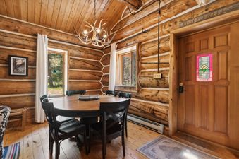 The dining area at Apres Chalet features a round wooden table, black chairs, log cabin walls, and an antler chandelier, with windows offering an exterior view.