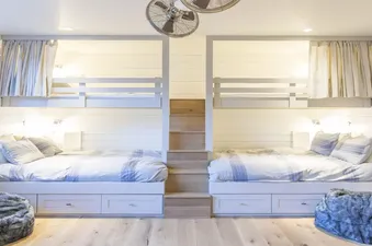 A bright bunk room at Overlook Haus with two sets of built-in bunk beds, each featuring a lower bed with storage drawers and an upper bunk, separated by a central wooden staircase.