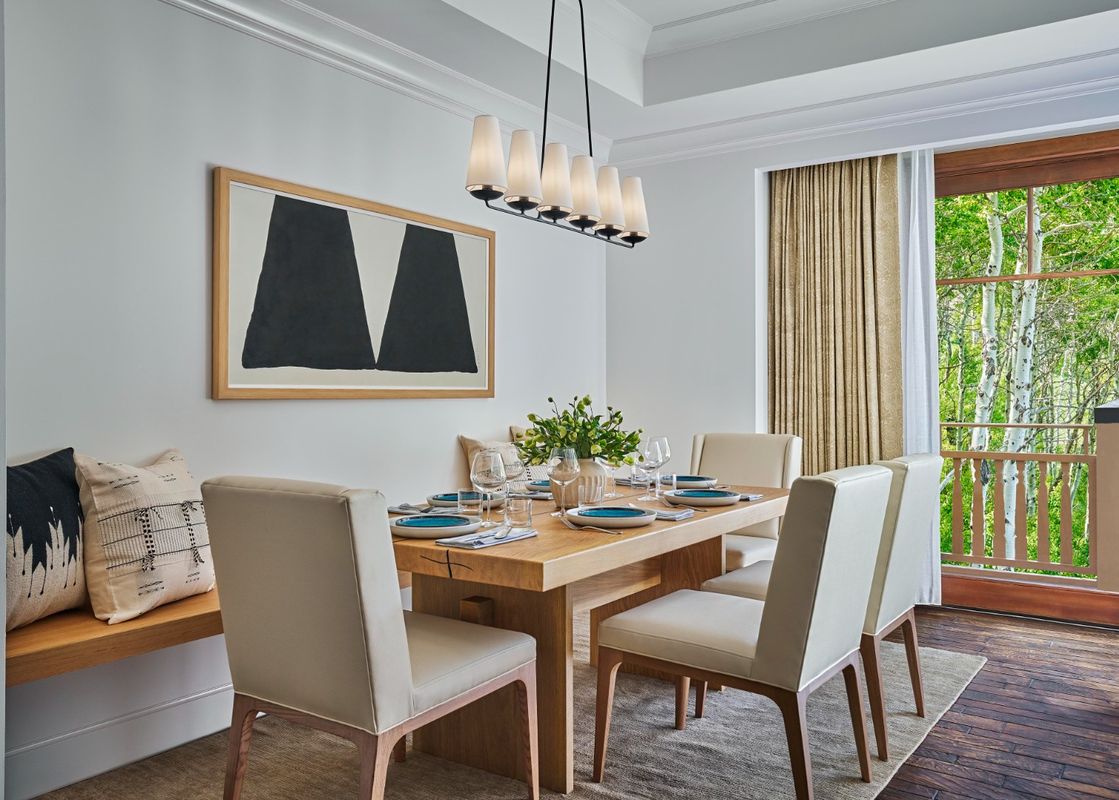 A modern dining area with a long wooden table set for six, featuring light-colored chairs, a large abstract painting on the wall, and a view of lush green trees outside a large window.