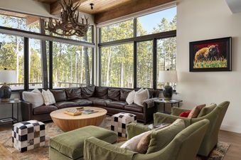 A rustic yet luxurious living room with a large leather sectional, round coffee table, and forest views through expansive windows.