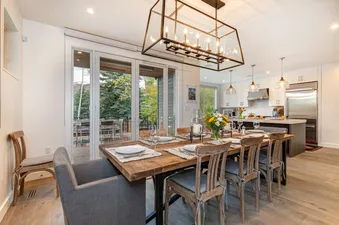 A dining room with a large wooden table and chairs, facing a patio with a view of the mountains and trees.