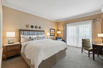A hotel bedroom with a king-sized bed, two bedside tables with lamps, a blue armchair, a desk with a green chair, and a large window with sheer and blackout curtains.