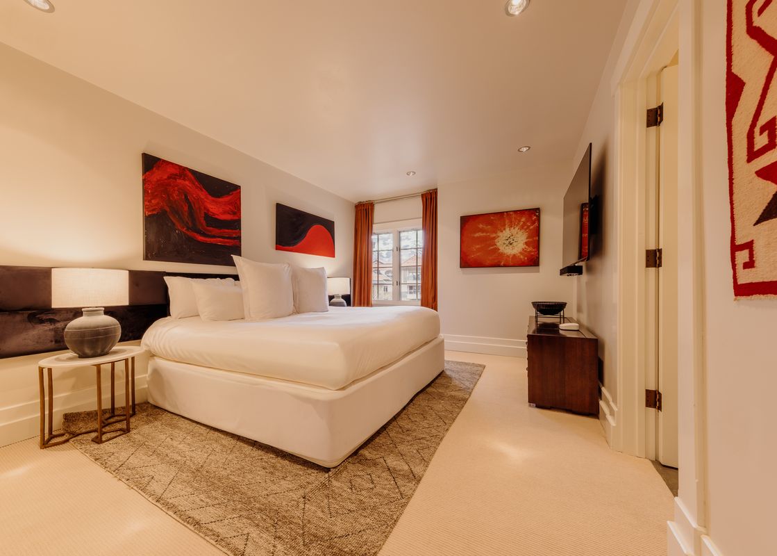 A serene bedroom at High West at the Plaza featuring a king-sized bed with white linens, artistic red and black abstract paintings, a modern bedside lamp, and a large window with orange curtains.
