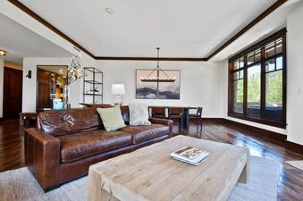 The comfortable living area of Arrowleaf Lodge 107 features a brown leather sofa, a wooden coffee table, and an adjacent dining area, with large windows offering a view of the outdoors.