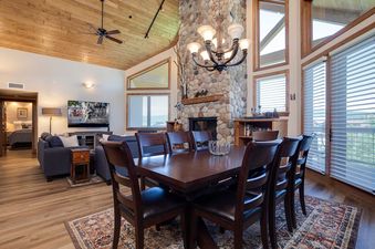 A luxurious living area at Chateau Chamonix CX343 featuring a stone fireplace, a cozy sofa, a large TV, and a dining table with elegant chairs, all bathed in natural light.
