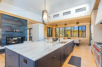 A luxurious kitchen at The Summit with a large marble island, a dark metal fireplace, and expansive windows showcasing an autumn forest view.
